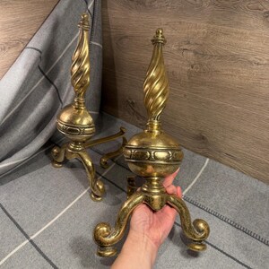 Pode incluir: Dois cães de fogo antigos de latão. Cada um apresenta um design de coluna torcida, uma esfera decorativa e três pernas curvas. Os cães de fogo têm um acabamento dourado e são exibidos em um cobertor listrado cinza e branco.