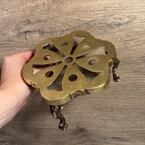 May include: A brass trivet with a floral design. The trivet has a flower-like shape with cut-out details and three decorative legs. The brass has a warm, golden hue, and the trivet is designed to protect surfaces from hot items.