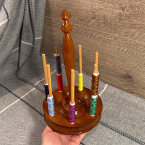 May include: A wooden thread spool holder with a central vertical post and multiple wooden pegs. The holder is filled with spools of colorful thread in various colors, including blue, red, yellow, and green. The holder is made of polished wood.