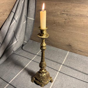 May include: A gold metal candlestick with ornate details and a single white candle.