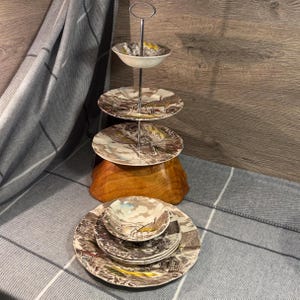 May include: A vintage tiered cake stand with three plates and a bowl, decorated with a landscape scene in brown, beige, and yellow. The stand is metal and wood, with additional matching plates and bowls.