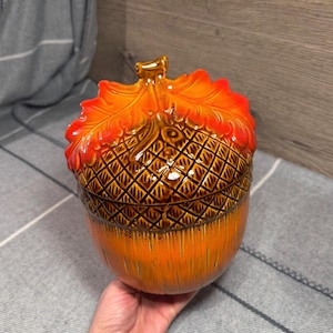 Peut inclure: Un pot en céramique en forme de gland avec un couvercle amovible. Le couvercle présente un capuchon marron texturé à motifs croisés et une feuille orange vif sur le dessus. La base du pot est orange brillant avec des détails brossés marron.