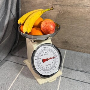 May include: A cream-coloured mechanical kitchen scale with a round white dial featuring black numbers and a red needle. A stainless steel bowl sits on top, filled with two yellow bananas, two orange oranges, and one red apple with visible text.