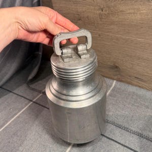 May include: A cylindrical aluminum canister with a brushed metallic finish. The container features a threaded neck and a hinged, rectangular-shaped handle attached to the top lid. The design is utilitarian, showcasing a smooth, unadorned surface.