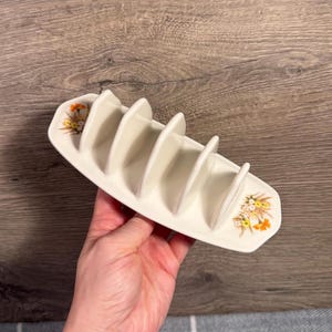 Puede incluir: Tostador de cerámica color crema con seis ranuras. El soporte tiene forma rectangular con extremos redondeados, decorado con motivos florales en amarillo, naranja y verde. Diseñado para sostener las tostadas en posición vertical. Mide unos 18 cm de largo.