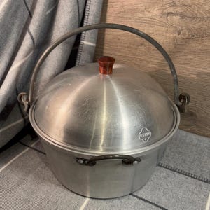 Può includere: Una pentola da cucina vintage in alluminio con coperchio e manico. La pentola ha una forma arrotondata con un pomello in legno sul coperchio. Il manico è in metallo e si incurva verso l'alto. La pentola è su una superficie grigia e bianca.