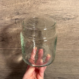 Könnte beinhalten: Ein klares Glas mit Deckel. Das Glas hat eine strukturierte Oberfläche und wird von einer Person gehalten. Das Glas ist leer und scheint sauber zu sein. Das Glas eignet sich zur Aufbewahrung von Lebensmitteln oder anderen kleinen Gegenständen.