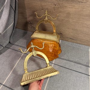 Puede incluir: Dos soportes para herramientas de chimenea de latón a juego con un diseño de arco semicircular, una base estriada y brazos curvos decorativos. Los objetos de tono dorado metálico tienen un acabado pulido y una forma estructurada.