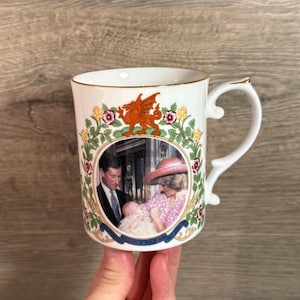 Puede incluir: Taza de cerámica blanca con borde y asa dorados. La taza presenta una imagen central de un retrato de la familia real, rodeada de un diseño floral y un emblema de dragón rojo. El asa tiene una forma decorativa.