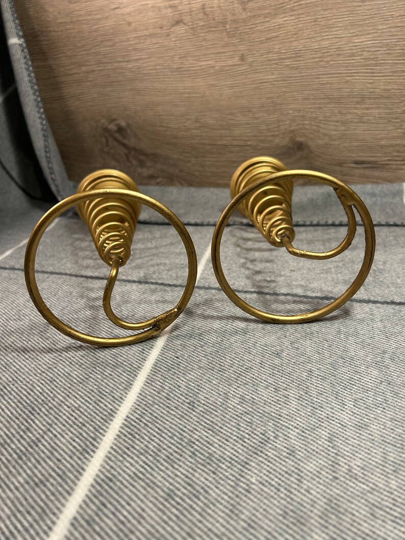 Peut inclure: Deux bougeoirs en m&eacute;tal dor&eacute; avec un design en spirale. Les bougeoirs sont sur une surface ray&eacute;e gris et blanc.