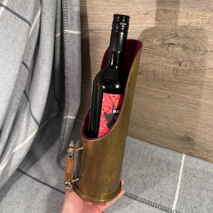 Puede incluir: Un portabotellas de vino de latón con asa e interior burdeos. Dentro del soporte hay una botella de vino tinto con una etiqueta roja y blanca. El asa tiene un mango de madera.