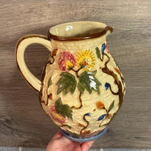 Könnte beinhalten: Eine antike Keramikkanne mit Blumenmuster. Die Kanne ist cremefarben mit braunen, grünen, gelben, rosa und blauen Akzenten. Das Blumenmuster zeigt Blätter, Ranken und Blumen.