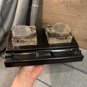 May include: A black inkwell with two glass inkwells featuring a grid pattern on the top. The inkwell is rectangular in shape and has a raised edge.