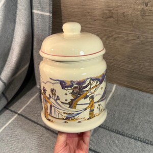 Puede incluir: Tarro de cerámica color crema con tapa, con una escena pintada a mano de personas cosechando. El tarro tiene un borde rojo en la tapa y una forma redondeada. La obra de arte representa figuras en un campo con un árbol.