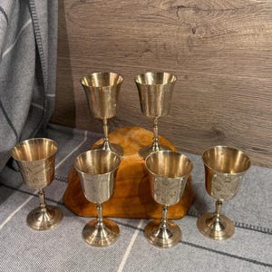May include: Six vintage brass goblets with ornate floral designs. The goblets have a classic chalice shape, with a round base and a slender stem. The goblets are arranged on a wooden surface and a gray and white striped cloth.