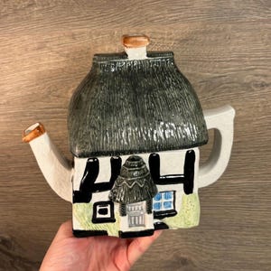 May include: A white ceramic teapot shaped like a cottage with a thatched roof. The teapot has black accents and a green lawn. The spout is shaped like a chimney.