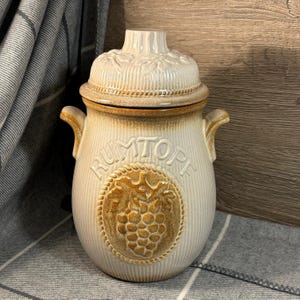 May include: A white ceramic jar with a brown lid and a brown grape design. The jar has the word "Rumtopf" written on it.