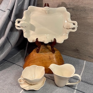 May include: A cream-colored serving tray with ornate handles and decorative accents, displayed on a wooden stand. A matching teacup and saucer, along with a creamer, are also visible, suggesting a tea set.