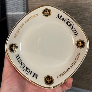 Może przedstawiać: Biały, kwadratowy ceramiczny talerzyk ze złotym obramowaniem. Na talerzyku widnieje napis "The Real Mackenzie Scotch Whisky" w czarnym kolorze. Obecne są również złote emblematy z motywem jelenia.