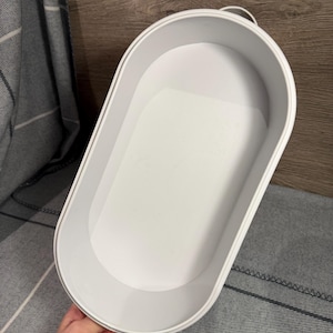 May include: A white oval-shaped storage tray with high, smooth walls and a flat base. The container features a minimalist design with a clean, matte finish and a small, integrated handle loop at one end, providing a simple and functional storage solution.