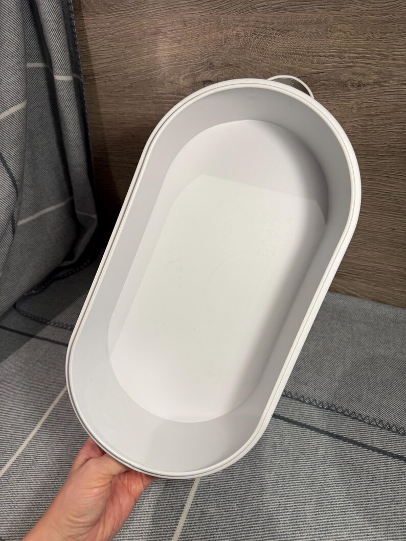 May include: A white oval-shaped storage tray with high, smooth walls and a flat base. The container features a minimalist design with a clean, matte finish and a small, integrated handle loop at one end, providing a simple and functional storage solution.