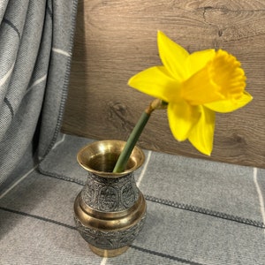 K&ouml;nnte beinhalten: Eine gelbe Narzisse in einer kleinen, verzierten Messingvase mit aufwendigen Designs. Die Vase steht auf einer grauen und wei&szlig;en gestreiften Decke.