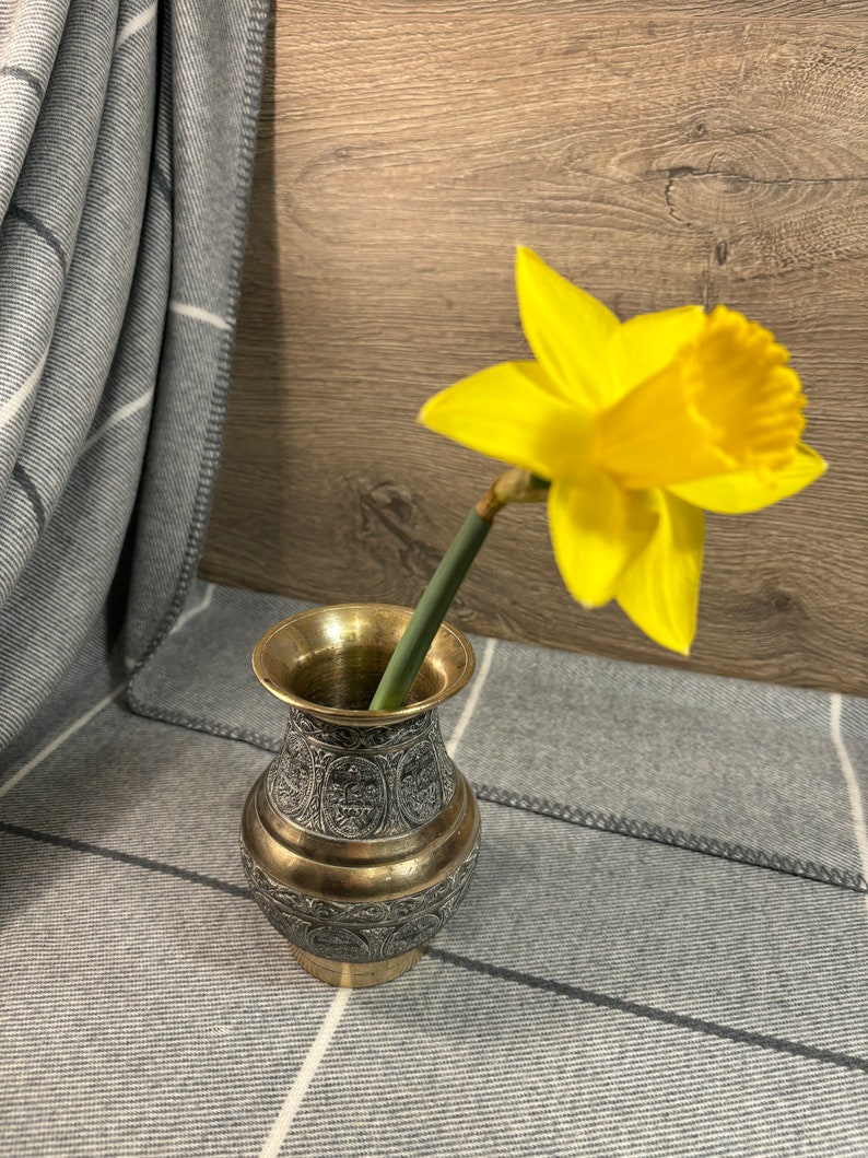 K&ouml;nnte beinhalten: Eine gelbe Narzisse in einer kleinen, verzierten Messingvase mit aufwendigen Designs. Die Vase steht auf einer grauen und wei&szlig;en gestreiften Decke.