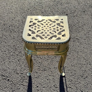 May include: A brass trivet with a decorative openwork design on the top. The trivet has curved legs and a detailed edge. The overall color is gold, and it is designed to protect surfaces from hot items.
