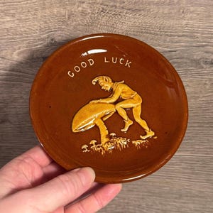 May include: A brown ceramic dish with the words "GOOD LUCK" in white lettering. The dish features a relief of a figure pushing a large mushroom. The figure and mushroom are in a lighter shade of brown.
