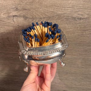 May include: A silver-toned metal container with a square opening and ornate, curved feet, filled with a cluster of wooden matches featuring dark blue tips. The container has a decorative, textured rim and a polished, reflective surface finish.