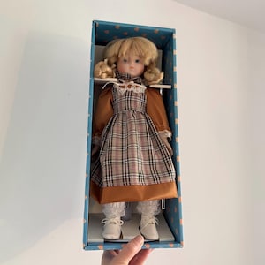 May include: A vintage doll with blonde pigtails, wearing a brown plaid dress with a white lace collar and cuffs, and white shoes. The doll is displayed in a blue box with a heart pattern.