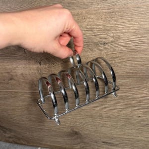 Puede incluir: Un tostador cromado con seis ranuras curvas. El soporte está hecho de metal brillante y tiene una base rectangular con pequeños pies. El soporte está diseñado para sostener la tostada en posición vertical.