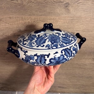 May include: A white and blue ceramic serving dish with a lid. The dish features a floral pattern in blue and has dark blue handles. The lid has a dark blue knob. The dish is oval-shaped and appears to be vintage.