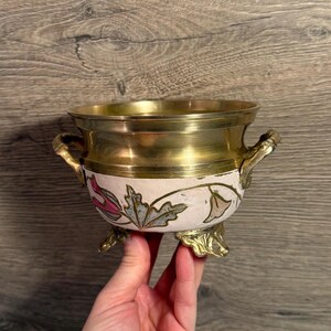 Könnte beinhalten: Kleine, dekorative Messingschale mit einem weiß lackierten Band mit floralen Designs in Rosa, Blau und Grün. Die Schale hat zwei Griffe und drei verzierte Füße. Innen und am Rand ist sie aus poliertem Messing.