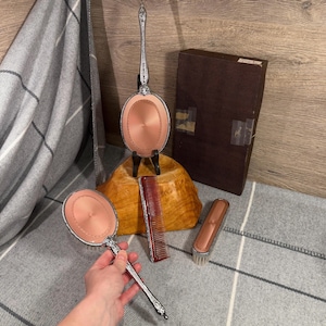 Könnte beinhalten: Ein Vintage-Frisier-Set mit einem Handspiegel, einem Standspiegel, einem Kamm und einer Bürste. Die Spiegel haben eine rosa Oberfläche und verzierte silberfarbene Griffe. Der Kamm ist rot und die Bürste hat eine rosa Oberfläche und weiße Borsten. Eine braune Box ist ebenfalls enthalten.