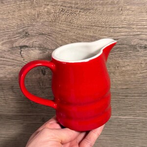 Può includere: Una piccola brocca in ceramica rossa con interno bianco. La brocca ha un manico curvo e un beccuccio per versare. L'esterno è rosso lucido e l'interno è bianco. La brocca è tenuta in una mano.