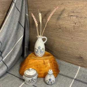 Könnte beinhalten: Eine graue Keramikvase mit Henkel, verziert mit blauen Blumen- und Vogelmotiven, hält getrocknete Weizenähren. Ein passendes Gefäß mit Deckel und ein Salzstreuer mit ähnlichen Designs stehen daneben. Die Artikel werden auf einem Holzsockel und einer grau karierten Decke präsentiert.