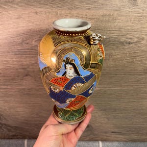 Könnte beinhalten: Handbemalte, antike japanische Vase mit detaillierter Illustration einer Frau in traditioneller Kleidung. Die Vase hat goldene, blaue, rote und grüne Akzente sowie eine kleine drachenartige Figur. Die Vase ist etwa 15 cm hoch.