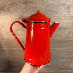 Puede incluir: Una cafetera de esmalte rojo brillante con tapa a juego y asa curva. La cafetera tiene un borde negro y un pico vertedor. El diseño recuerda a los utensilios de cocina vintage.