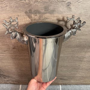 Puede incluir: Una cubitera de metal plateado con dos asas con forma de cabeza de ciervo. La cubitera tiene forma cilíndrica con un interior oscuro. Las cabezas de ciervo tienen cuernos y rasgos faciales detallados. La cubitera está sujeta por una mano.