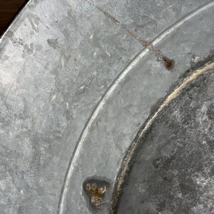 Op de afbeelding: Een close-up van een cirkelvormig gegalvaniseerd metalen oppervlak met een uitgesproken, kristallijne textuur. Het metaalgrijze materiaal vertoont zichtbare tekenen van oxidatie, waaronder kleine bruine roestvlekken en een dunne kras.