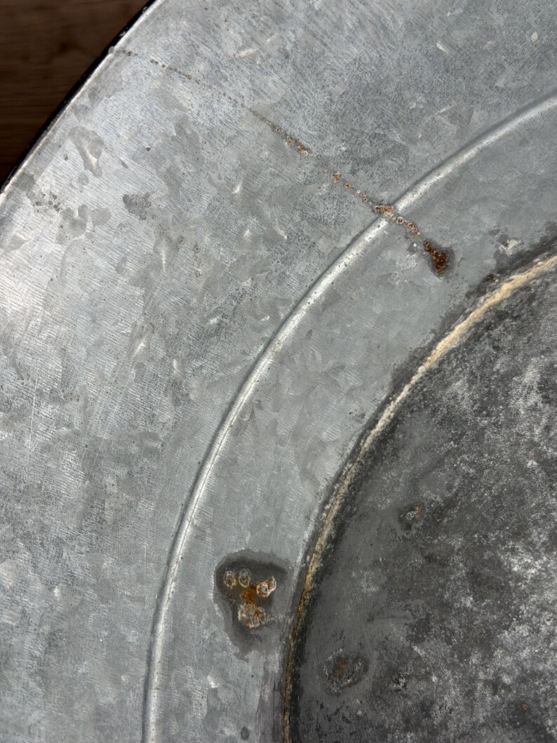 Op de afbeelding: Een close-up van een cirkelvormig gegalvaniseerd metalen oppervlak met een uitgesproken, kristallijne textuur. Het metaalgrijze materiaal vertoont zichtbare tekenen van oxidatie, waaronder kleine bruine roestvlekken en een dunne kras.