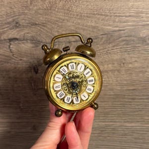 May include: A vintage brass alarm clock with a gold face and black Roman numerals. The clock has a handle and two bells on top. The clock is held in a hand.