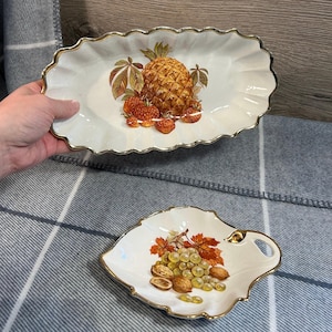 Może przedstawiać: Dwa vintage ceramiczne talerze do serwowania z pozłacanymi krawędziami. Większy talerz jest owalny i przedstawia ananasa, truskawki i liście. Mniejszy talerz ma kształt liścia i przedstawia winogrona, orzechy i liście.