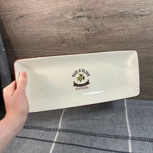 Può includere: Piatto da portata rettangolare in ceramica bianco sporco con bordo marrone. Il piatto presenta le parole "Olio d'Oliva" e "Buon Appetito Tutto Delizioso" con un disegno di ramo d'ulivo. Una mano tiene il piatto.