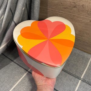 May include: Heart-shaped decorative box with a retro design. The lid features a vibrant pattern of orange, coral, and yellow heart-shaped petals radiating from the center. The box has a white base with pink and white stripes and a gold trim.