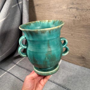 May include: A turquoise ceramic vase with a flared rim, a pedestal base, and two double-loop handles on each side. The glaze features a mottled finish with darker green and brown tones blending into the vibrant turquoise body of the vessel.