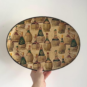 Op de afbeelding: Ovale serveerschaal met een herhalend patroon van wijnflessen in beige, groen en rood. De schaal heeft een zwarte rand. De flessen zijn afgebeeld met stro-achtige bekledingen. De schaal wordt door een hand tegen een witte achtergrond gehouden.