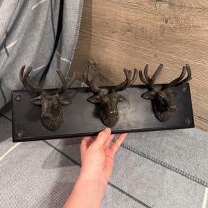 Puede incluir: Un perchero de pared rectangular negro con tres ganchos en forma de cabeza de ciervo en tono bronce oscuro. Cada cabeza de ciervo está esculpida con astas y rasgos faciales detallados. La base es un panel negro con orificios para tornillos.