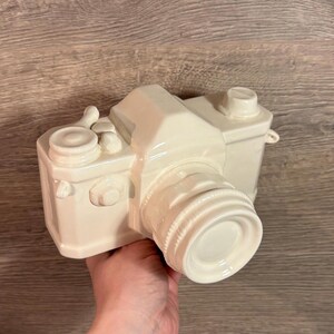 May include: A white ceramic camera with a lens and a few buttons. The camera is a decorative object.
